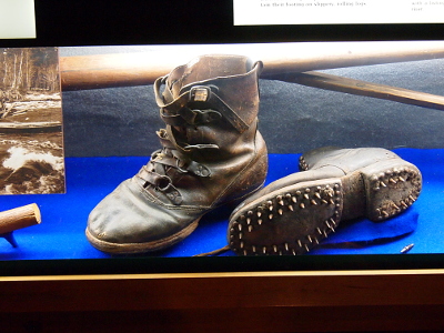 [A pair of boots in a case with one boot upright making the buckles visible while the other lies on its side showing the three rows of spikes around the perimeter of the top of the boot and around the heel.]